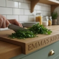 Here is the descriptive alt text for your generated lifestyle image, designed to help with SEO and web accessibility: "A handcrafted solid oak worktop saver with a front-edge stability lip, featuring custom 'Avril & Jim' engraving with hen icons, shown in a modern sage green kitchen with white subway tile backsplash and fresh herbs being prepared.