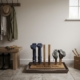 A wooden boot holder with vertical pegs stands against a light beige wall in a rustic boot room. The base of the holder is engraved with the text 'Rita's Mucky Boots.' Two navy blue wellington boots and a pair of hiking shoes are stored upside down on the pegs. The boot holder sits inside a stone-colored drip tray on a tiled floor. In the background, there is a wooden bench with gloves, a wall-mounted coat rack with jackets and a hat, and a wooden crate containing gardening tools next to a bright window.
