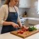 A woman in a denim apron slicing tomatoes on a personalized solid oak chopping board engraved with "Ryan’s Kitchen" in a bright modern kitchen.