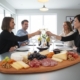 A high-angle, wide shot of a modern dining room where four friends are gathered around a sleek grey table, enjoying a social gathering. In the foreground, a large, oval-shaped oak wood serving board is laden with an abundant spread of cheese and charcuterie, including brie, hard cheeses, salami, prosciutto, grapes, walnuts, and dried apricots. In the background, two men and two women are smiling and raising their glasses of white wine in a celebratory toast. The room is bright and airy with minimalist decor, featuring large windows and a contemporary pendant light overhead.