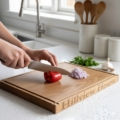 A high-quality, solid oak chopping board sits on a bright, modern white countertop. The board features a deep juice groove around the edge and the personalized engraving "Hillview House" on the front side. A person is shown slicing a vibrant red bell pepper with a chef's knife, next to a pile of diced shallots, a head of garlic, and fresh parsley. The background shows a clean kitchen with a stainless steel faucet, white ceramic canisters, and wooden utensils.