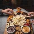 A large wooden snack board filled with popcorn, pretzels, cookies, and candy, centered between two people reaching for treats during a movie night.