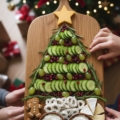 A wooden charcuterie board featuring a Christmas tree design made of sliced cucumbers, grapes, pomegranate seeds, and rosemary, accented with a cheese star and festive snacks.