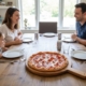 Artisan handcrafted round oak pizza board with a handle, holding a flatbread with green herb toppings on a speckled white kitchen countertop.