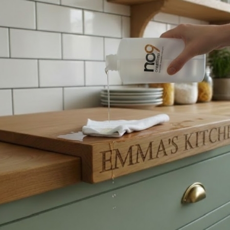 A person’s hand pours clear mineral oil from a semi-transparent plastic bottle onto a white cloth resting on a thick wooden cutting board. The bottle features a modern black and wood-textured logo that reads "no9 creations." The cutting board, which is engraved with the words "EMMA'S KITCHEN" on its side, is positioned on a light oak countertop in a bright kitchen with green cabinetry and white subway tile backsplashes.